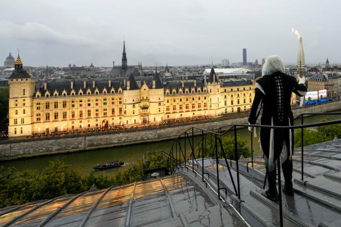 A torchbearer carries the Olympic flame over a building along the Seine River in Paris, France, during the opening ceremony for the 2024 Summer Olympics, Friday, July 26, 2024. (AP Photo/Bernat Armangue, Pool)