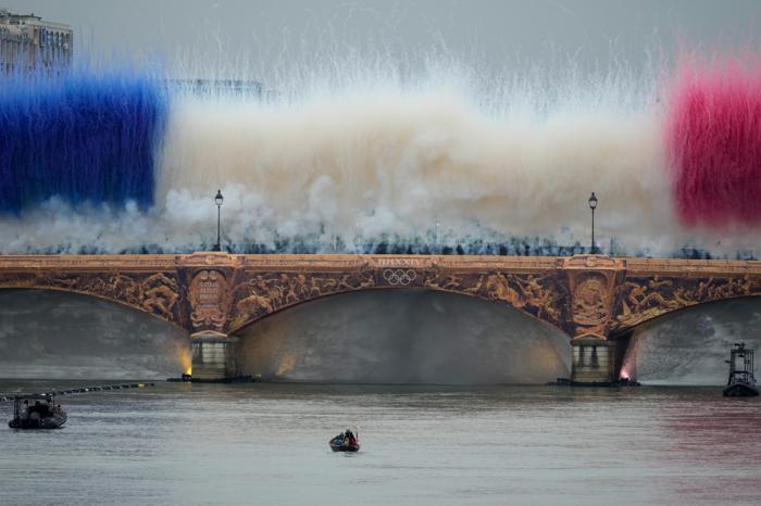 The Olympic torch travels by boat as ceremonial smoke in the colors of the France flag appear over the Seine River Paris, France, during the opening ceremony of the 2024 Summer Olympics, Friday, July 26, 2024. (AP Photo/Matthias Schrader)