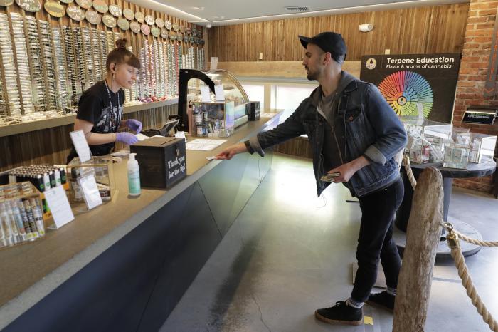 In this photo taken March 24, 2020, Christopher Ellis, right, leans in to show his identification to budtender Maegan Clark, left, as he stands on a line taped on the floor to designate a social distancing separation due to the new coronavirus, while shopping at The Reef Capitol Hill, a marijuana store in Seattle.