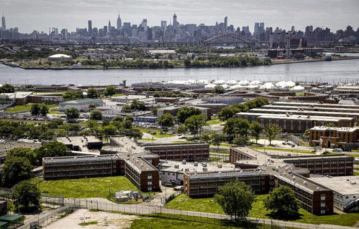 The Rikers Island jail complex in the foreground within the East River and the New York skyline in the background.