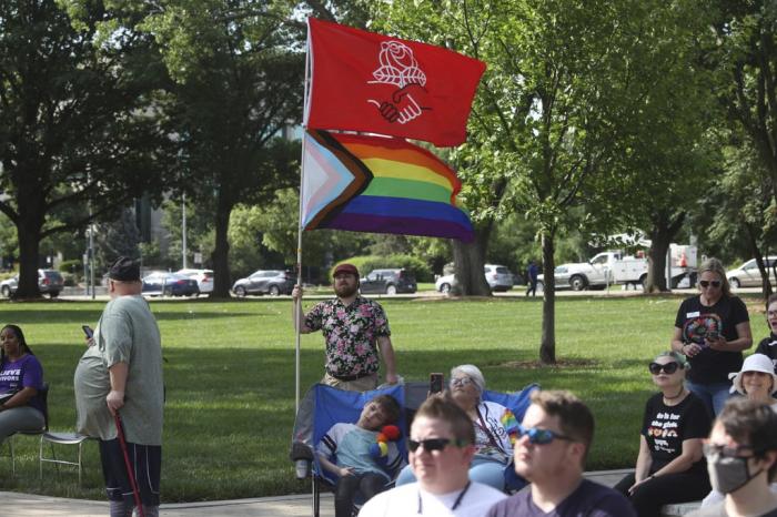 Kirby Evers, a 31-year-old bisexual Lawrence, Kan., resident, displays the red flag of the Democratic Socialists of America and a Pride flag during a rally outside the Kansas Statehouse in favor of transgender rights, Friday, June 30, 2023, in Topeka, Kan. 